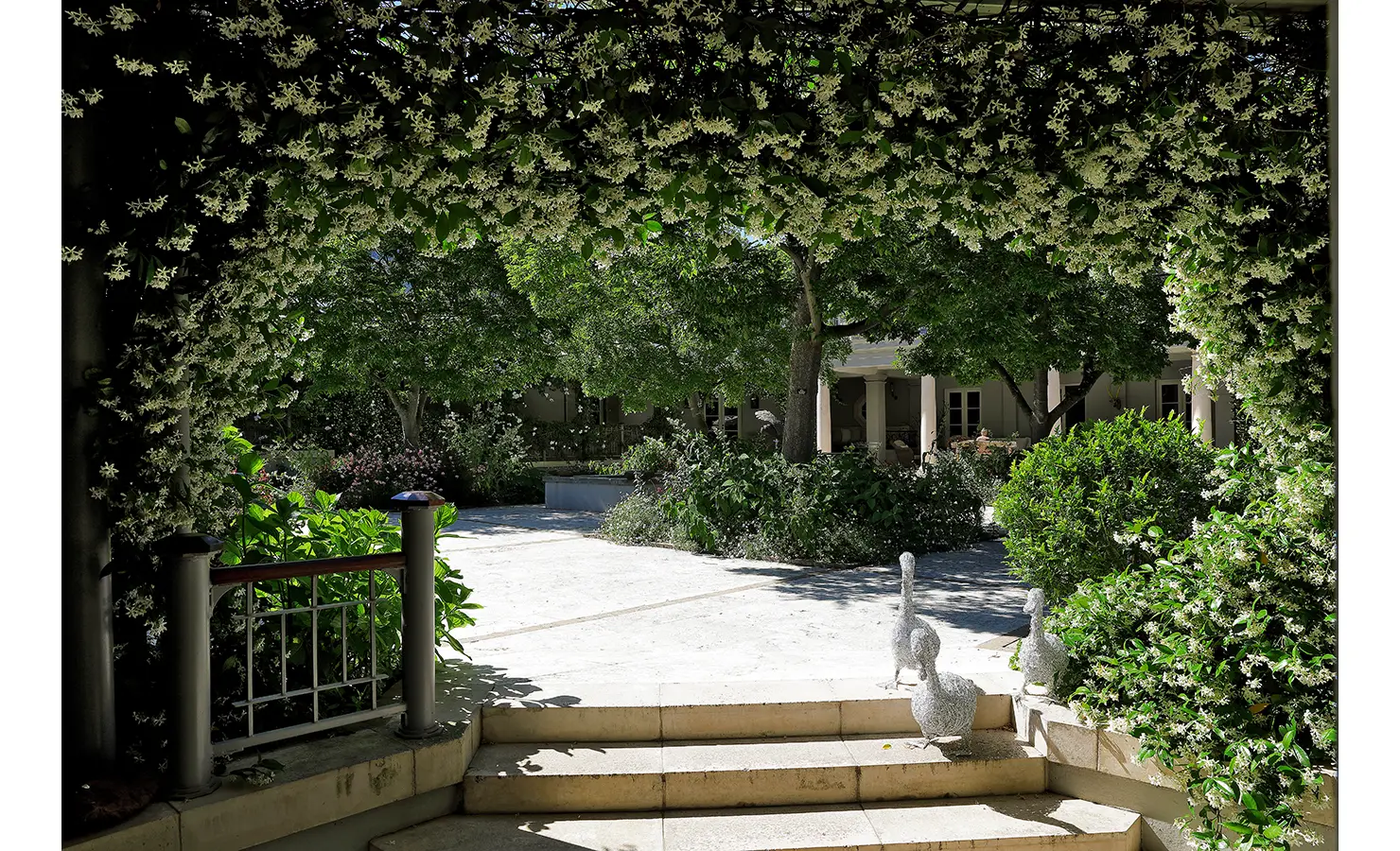 Perfect Hideaways shaded courtyard framed by an arch of white flowering vines, leading to a serene garden with sculptures and lush greenery, Franschhoek, Varuna.
