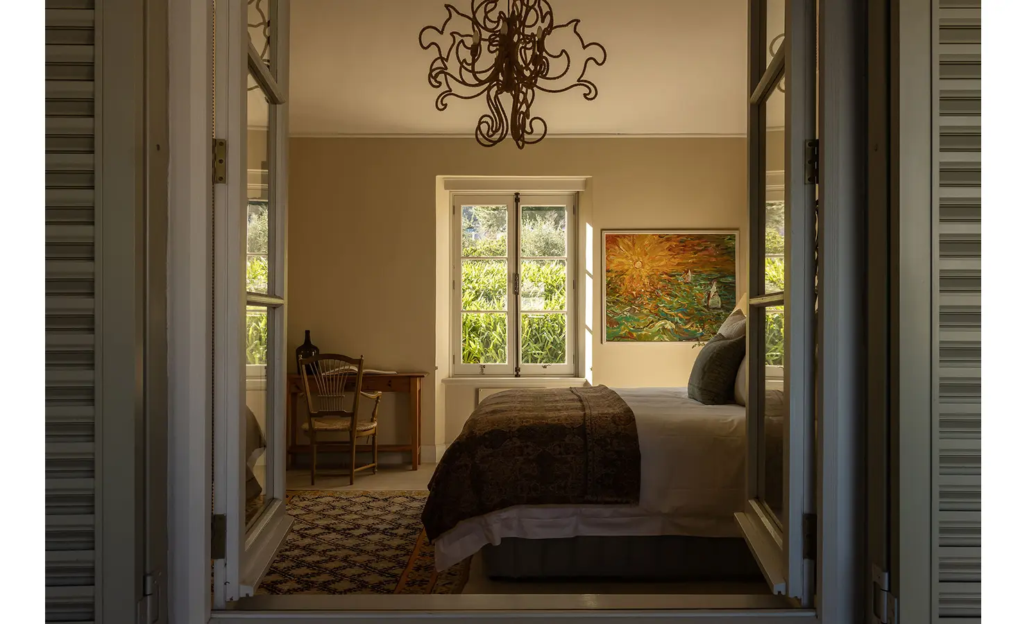 Perfect Hideaways cozy bedroom with warm lighting, a sun-themed artwork, and a desk beside a window overlooking the garden, Franschhoek, Varuna.