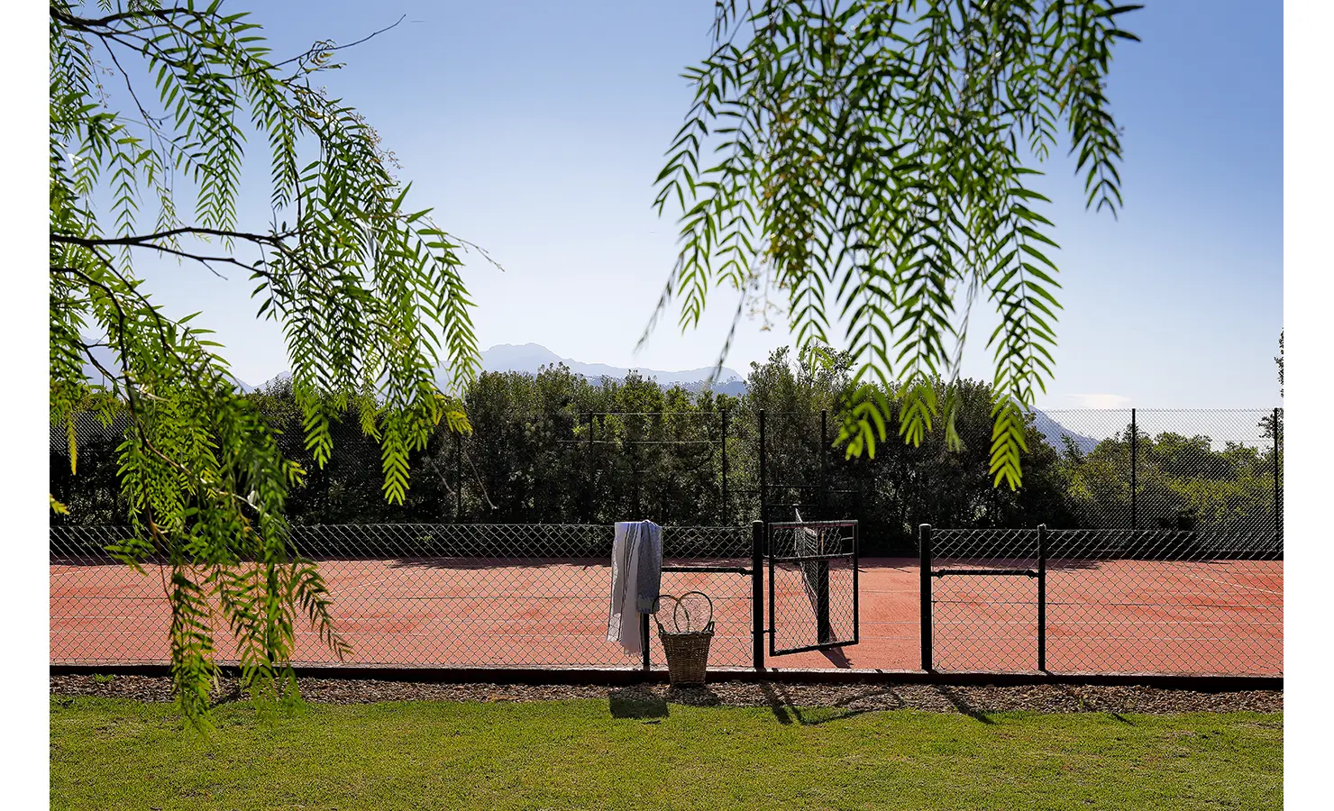 Perfect Hideaways sunlit tennis court bordered by trees and mountain views, with a basket of rackets and towels hanging by the gate, Franschhoek, Varuna.