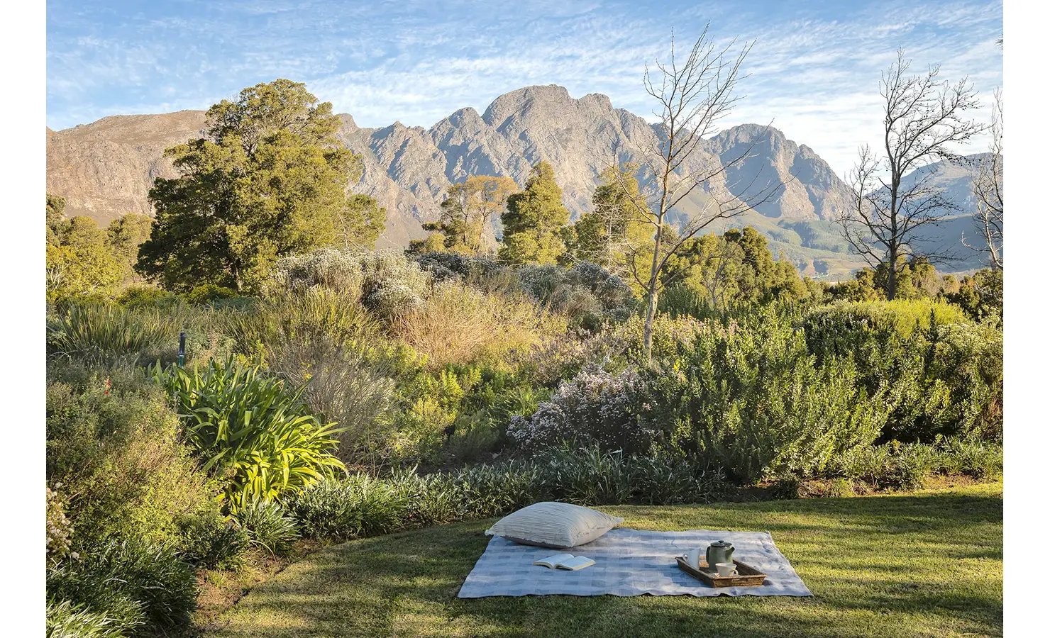 Perfect Hideaways tranquil garden picnic spot with a blanket, pillow, and tea set laid out amid lush vegetation and mountain vistas, Franschhoek, Varuna.