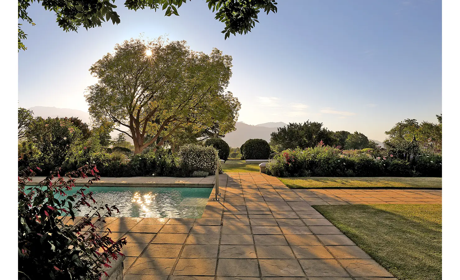 Perfect Hideaways golden sunset view over a manicured garden and poolside terrace, with sunlight filtering through trees and distant mountains, Franschhoek, Varuna.