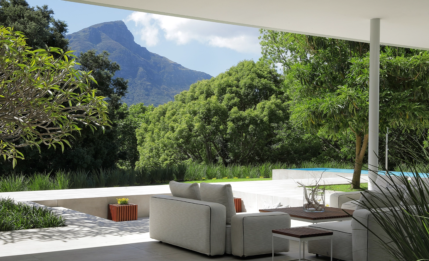 Perfect Hideaways A serene outdoor living space with modern white sofas arranged beneath a sleek overhang, opening onto a sunlit terrace surrounded by lush green trees, manicured planting, and a glimpse of a blue swimming pool, all framed by the dramatic mountain backdrop under a bright sky Pure Living, Bishopscourt