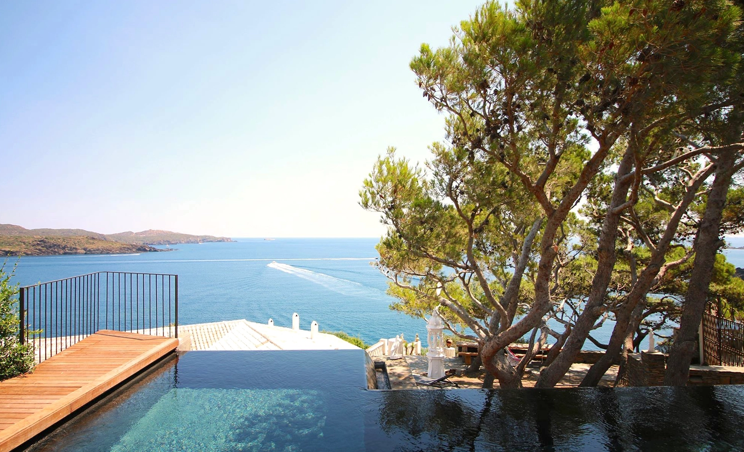 Perfect Hideaways: A serene coastal scene featuring an infinity pool overlooking calm, sparkling blue water, with rugged hills in the distance, a speedboat’s white wake cutting across the bay, and tall pine trees framing the view beside sunlit terraces and Mediterranean architecture, Cadaques, Casa Lligat.