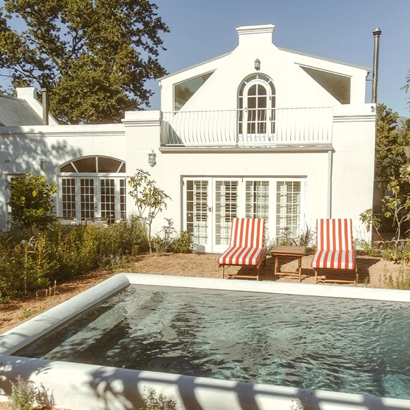 Perfect Hideaways A serene garden courtyard featuring a glistening rectangular plunge pool in the foreground, bordered by soft gravel and lush green plantings. Two red-and-white striped sun loungers with a small wooden side table sit facing the water, catching warm sunlight. The backdrop is a charming white Cape Dutch–inspired house with arched windows, French doors, and a balcony framed by mature trees, creating a peaceful, elegant retreat Franschhoek, Perla House