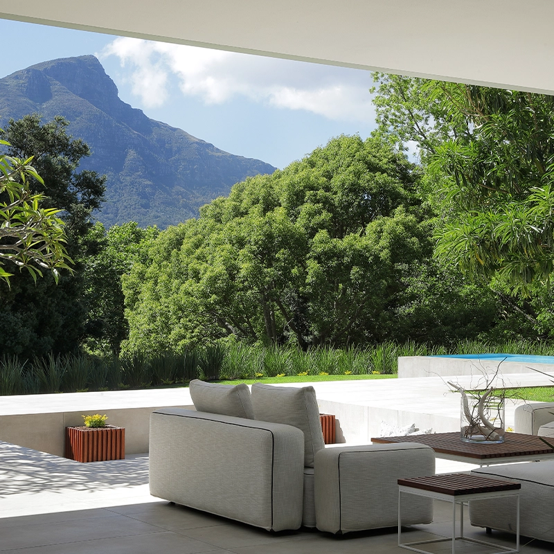 Perfect Hideaways A serene outdoor living space with modern white sofas arranged beneath a sleek overhang, opening onto a sunlit terrace surrounded by lush green trees, manicured planting, and a glimpse of a blue swimming pool, all framed by the dramatic mountain backdrop under a bright sky Pure Living, Bishopscourt