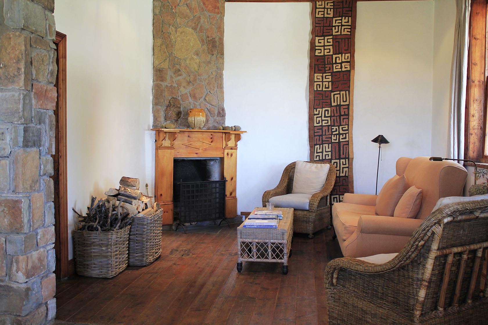 Perfect Hideaways- A cozy living room with rustic stone walls, a wooden fireplace surround, wicker armchairs, a soft peach-toned sofa, and woven baskets filled with firewood, creating a warm and inviting atmosphere. Karoo Cottage, Sneeuberg Nature Reserve, Eastern Cape, South Africa