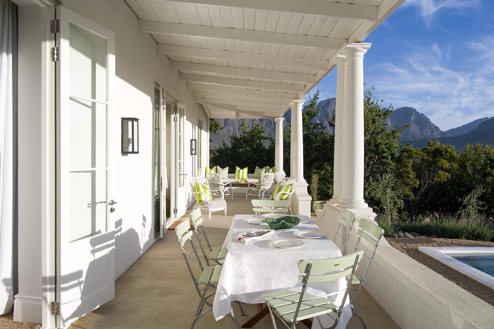 Perfect Hideaways – A long shaded veranda with white wooden beams and columns, set with a dining table draped in a white cloth and pale green folding chairs, opening toward a mountain view. At the far end, white wicker chairs with green-striped cushions create a relaxed seating area overlooking trees and distant peaks. La Cotte Orchard Cottages, Franschhoek South Africa