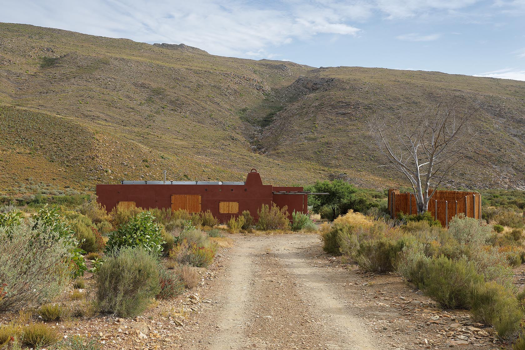Perfect Hideaways – A long gravel road leads toward a low, terracotta-coloured cottage nestled in a rugged Karoo valley, surrounded by soft green scrub and framed by steep, rocky hills under a pale blue sky. To the right, a circular wooden enclosure wraps around a leafless tree, adding a sculptural element to the wild landscape. Sheperd's Cottage, Prince Albert