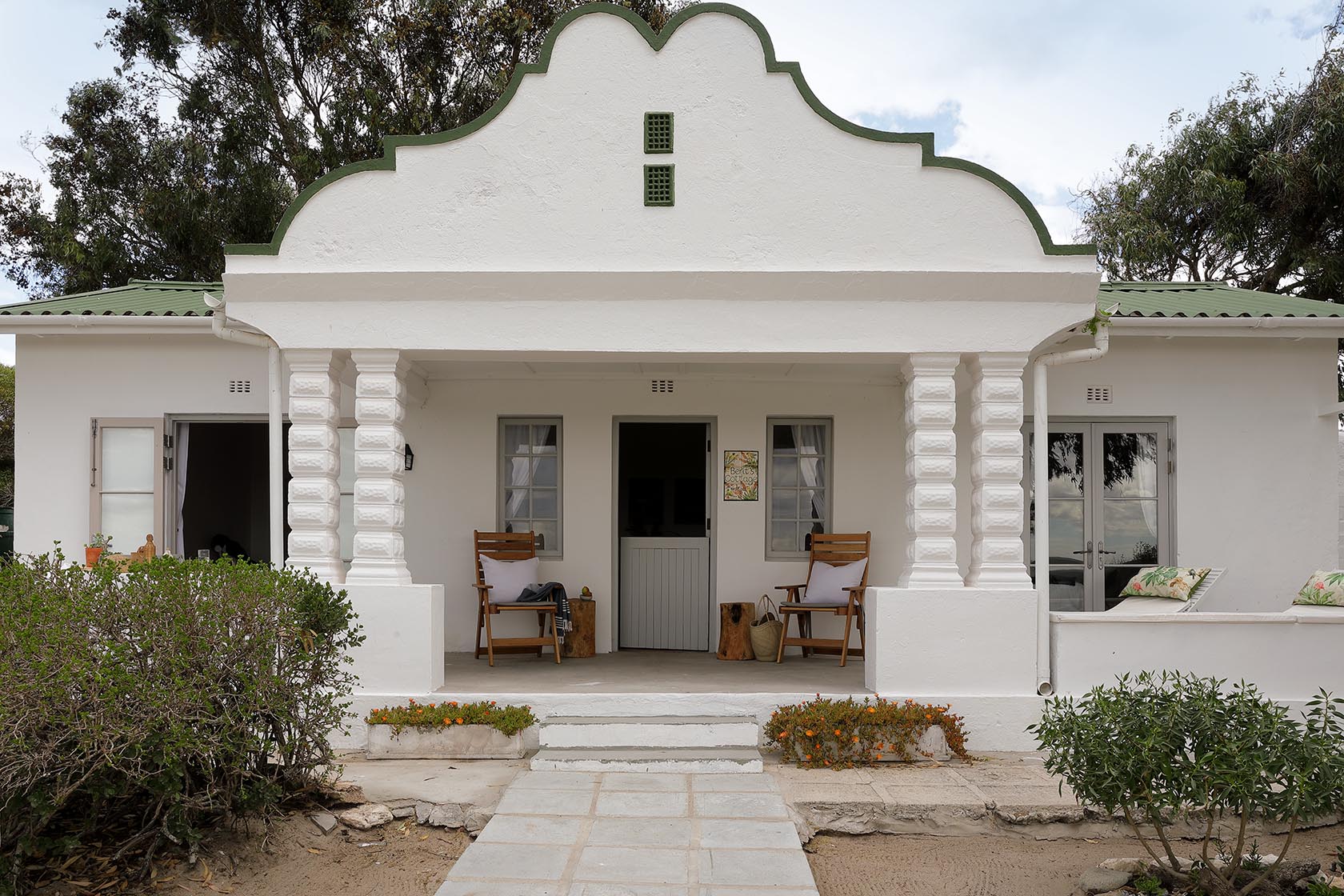 Perfect Hideaways – A charming whitewashed cottage with a classic Cape Dutch–inspired gable, green-trimmed roof, and a deep front veranda framed by sculpted pillars. Two wooden chairs with cushions sit on either side of the doorway, while simple garden beds and low shrubs border the sandy path leading to the entrance. Berit’s Cottage, Churchhaven, South Africa.