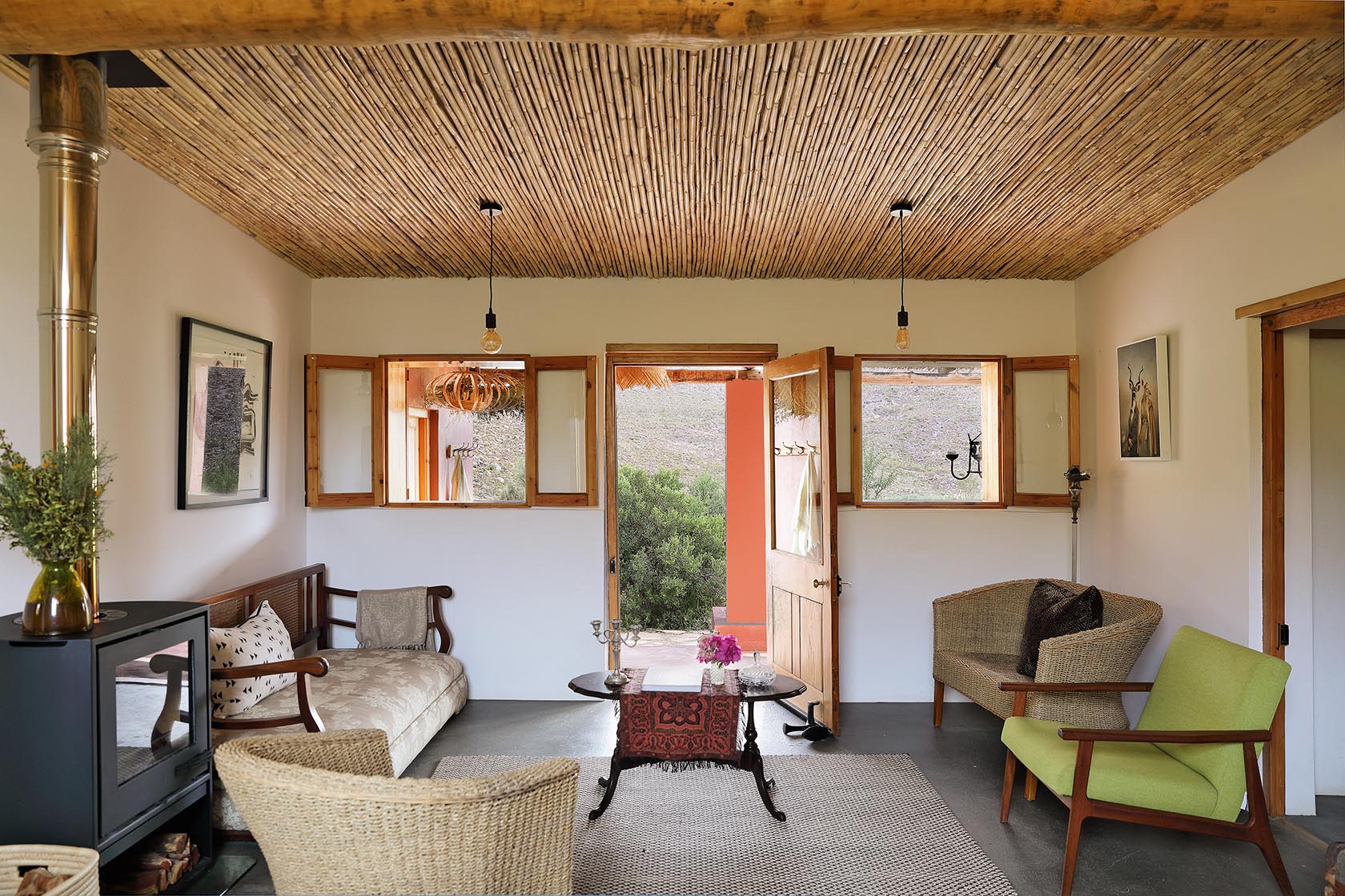 Perfect Hideaways – A cosy living room with a bamboo-lined ceiling and simple pendant lights features woven armchairs, a wooden daybed with patterned cushions, and a wood-burning fireplace. Sunlight enters through open windows and a door leading to the stoep, revealing the rocky hills beyond. Sheperd's Cottage, Prince Albert