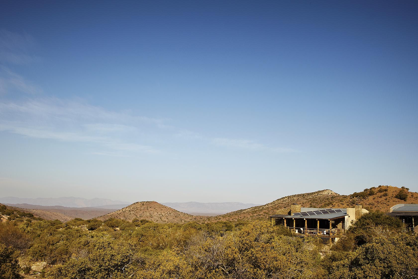 Perfect Hideaways–a vast Karoo landscape under a wide blue sky with rolling shrub-covered hills, featuring a secluded stone-and-timber cottage partially visible on the right, blending naturally into the surrounding wilderness–Stoney Cottage, Touwsberg Private Game and Nature Reserve, Western Cape, South Africa