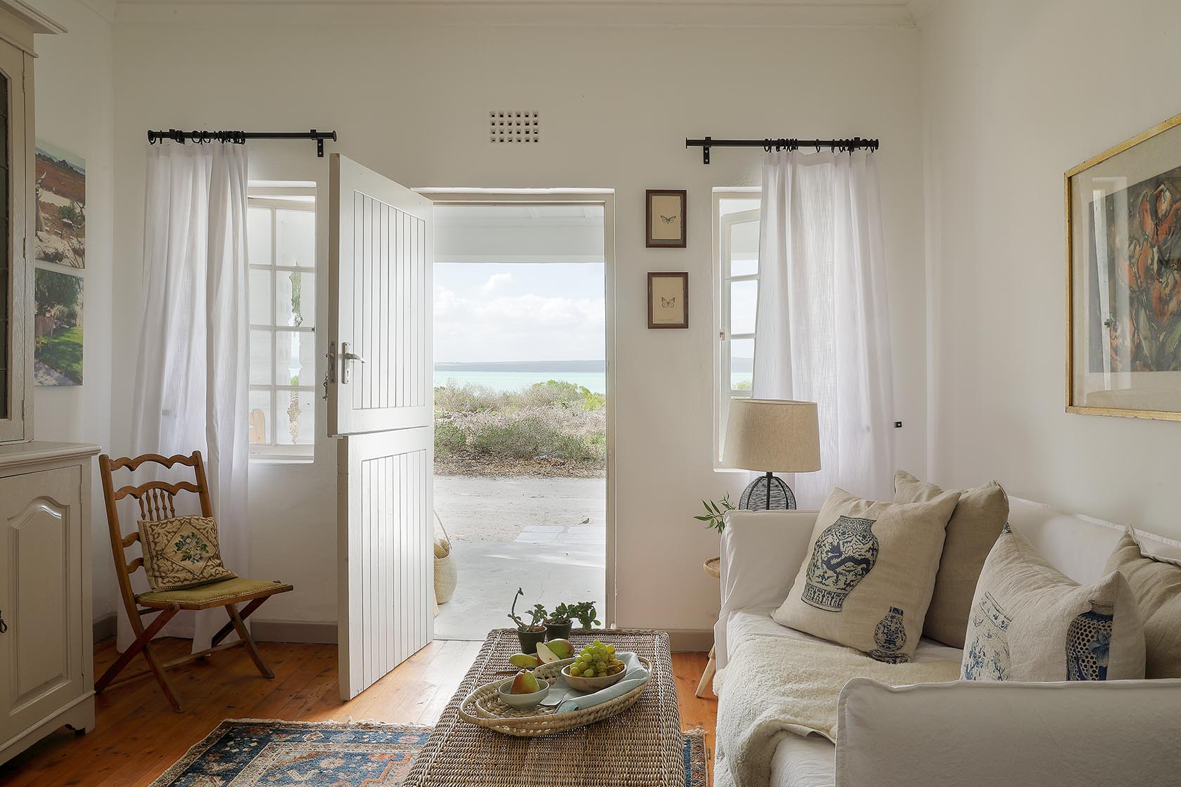 Perfect Hideaways – A bright, airy living space with white walls, sheer curtains, and a half-open stable door revealing views of dune shrubs and the lagoon beyond. A cozy white sofa with patterned cushions sits beside a woven coffee table arranged with fresh fruit, creating a serene, coastal atmosphere. Berit’s Cottage, Churchhaven, South Africa