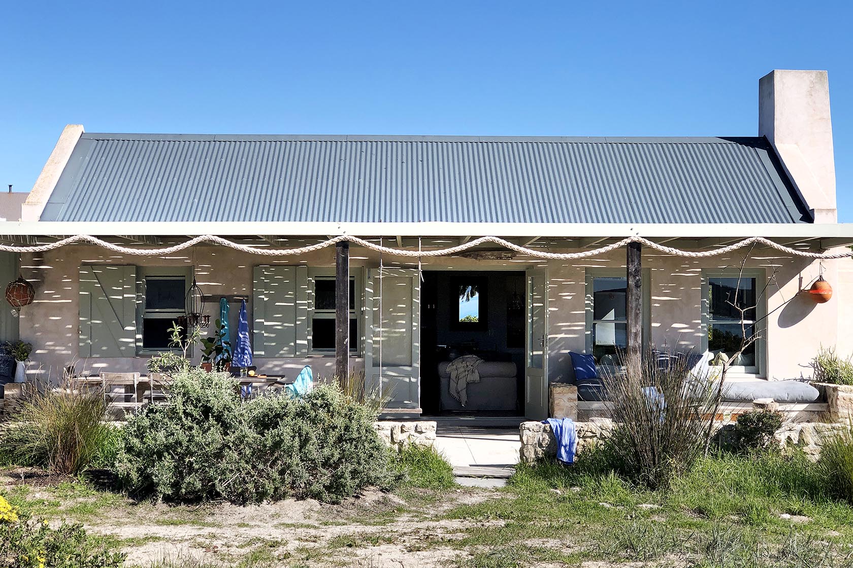 Perfect Hideaways- A closer view of the cottage façade, showing pastel shutters, a shaded veranda with seating areas, and wild coastal vegetation in the foreground beneath bright blue skies. Milkwood Cottage, Churchhaven, South Africa