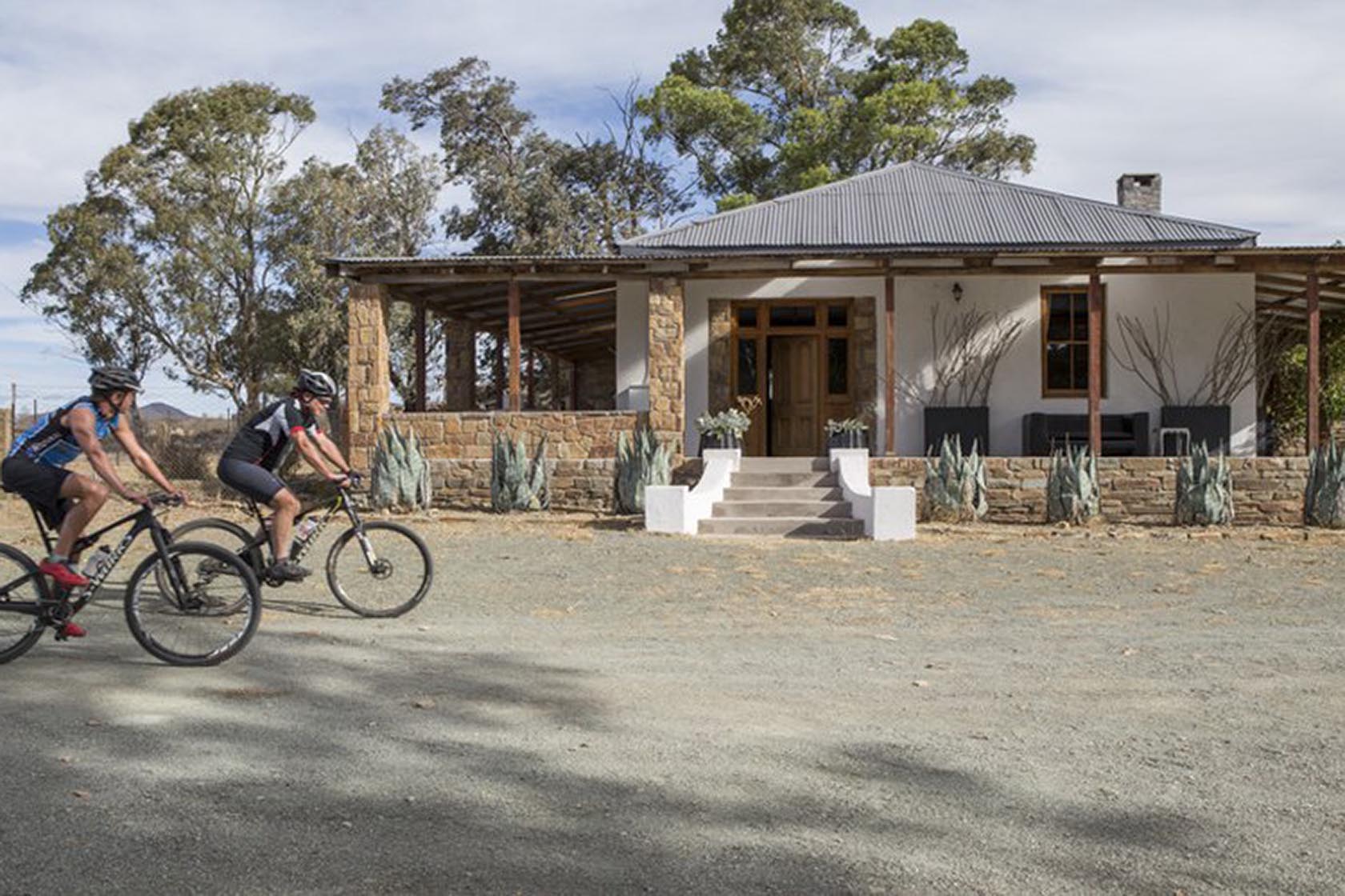 Perfect Hideaways- Two cyclists in helmets ride mountain bikes past the front of a rustic Karoo cottage, framed by stone pillars, agave plants, and tall trees under a bright sky. Karoo Cottage, Sneeuberg Nature Reserve, Eastern Cape, South Africa