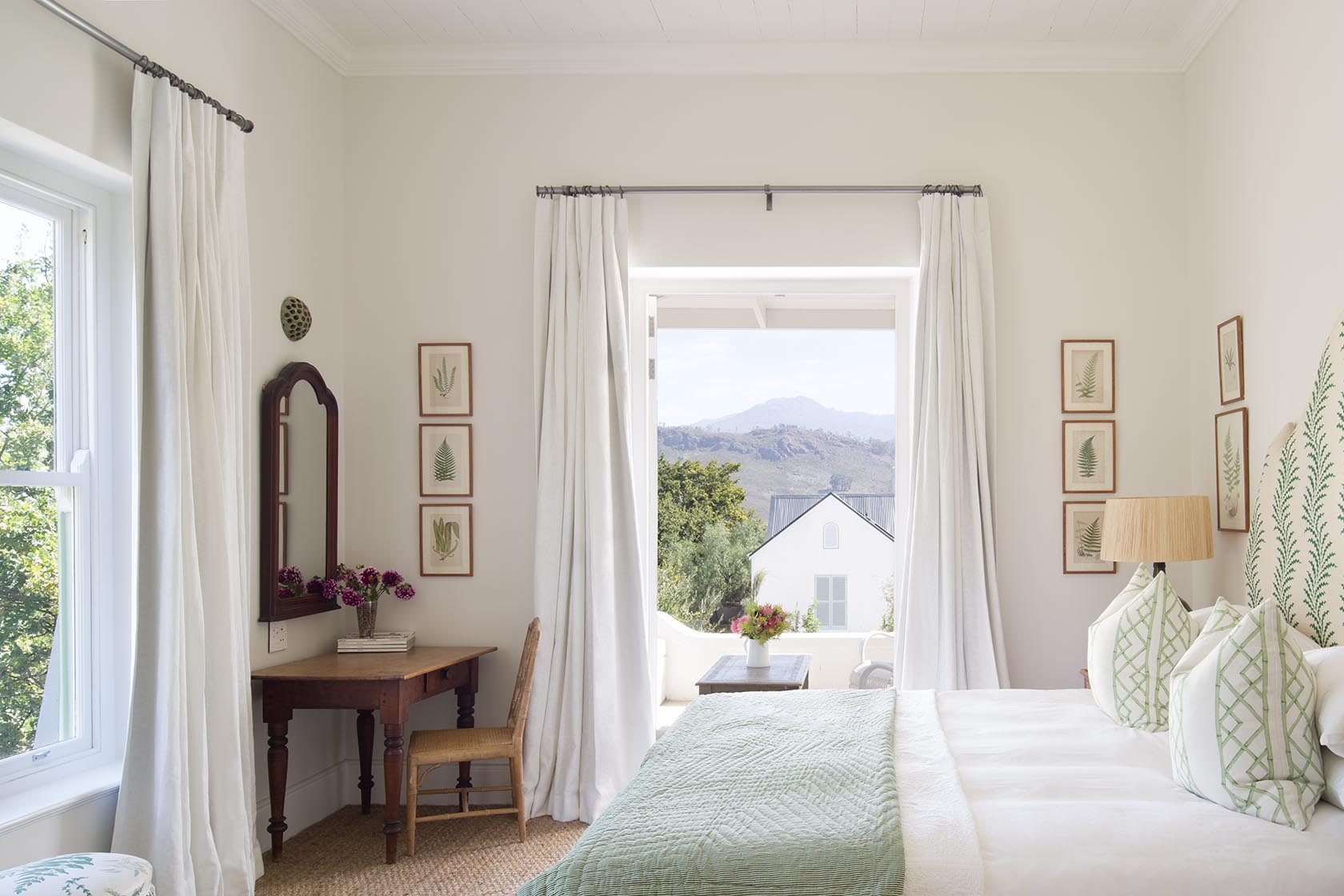 Perfect Hideaways – A bright bedroom with white curtains pulled aside to reveal a mountain view through open double doors, featuring a green-accented bed, botanical prints on the walls, and a small wooden writing desk with flowers, blending soft natural light with fresh, airy décor. La Cotte Orchard Cottages, Franschhoek South Africa