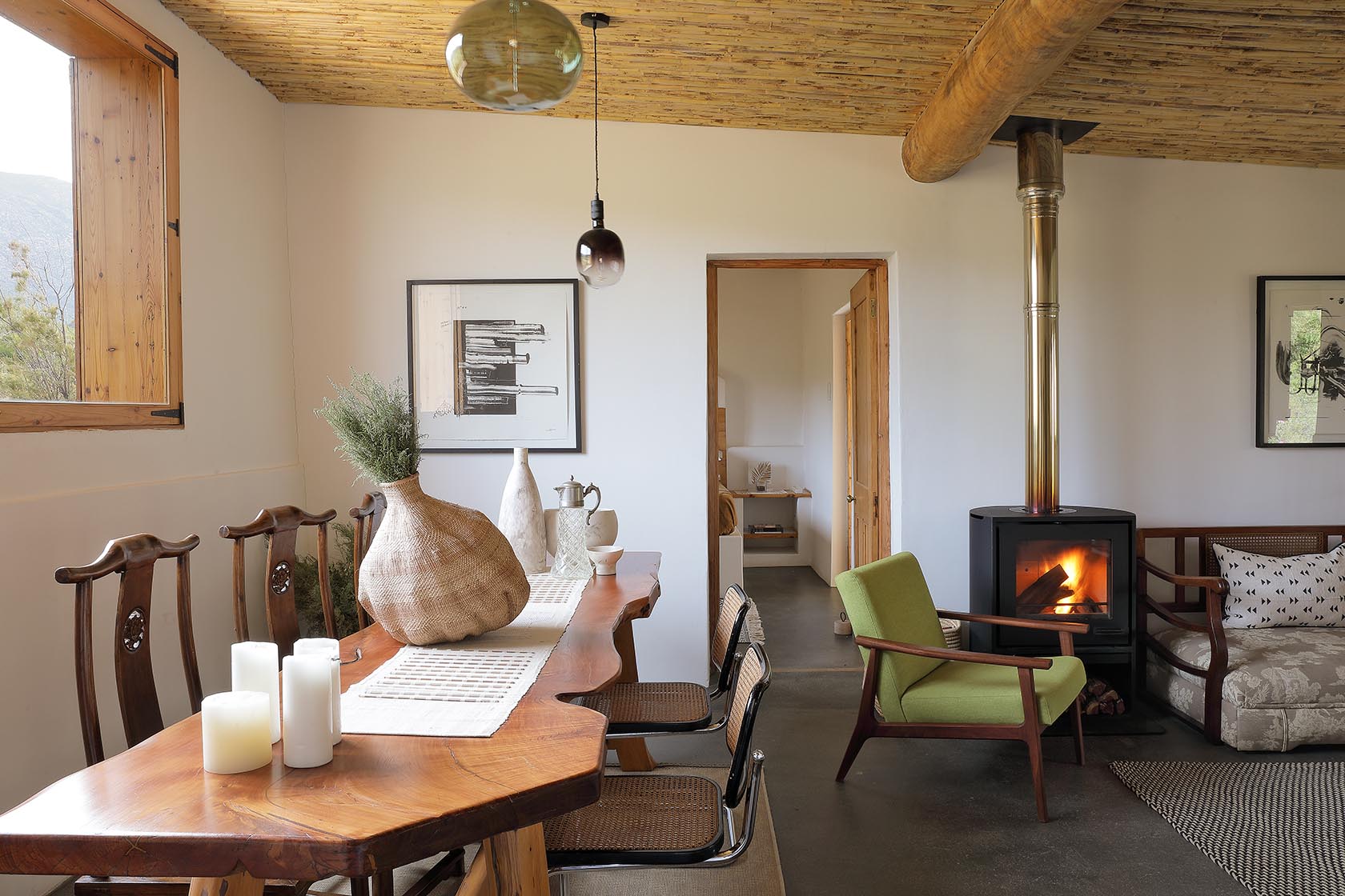 Perfect Hideaways – A warm and inviting open-plan dining and living area with a live-edge wooden dining table, wicker and wooden chairs, and a glowing fireplace. Natural textures, framed artwork, and soft furnishings create a calm, rustic-modern atmosphere. Sheperd's Cottage, Prince Albert
