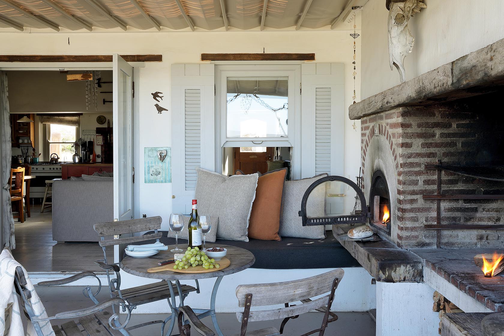 Perfect Hideaways–Cozy outdoor nook with cushions beside the brick pizza oven, a small round table set with wine and grapes, and open doors into the cottage. Church Cottage, Churchhaven, South Africa
