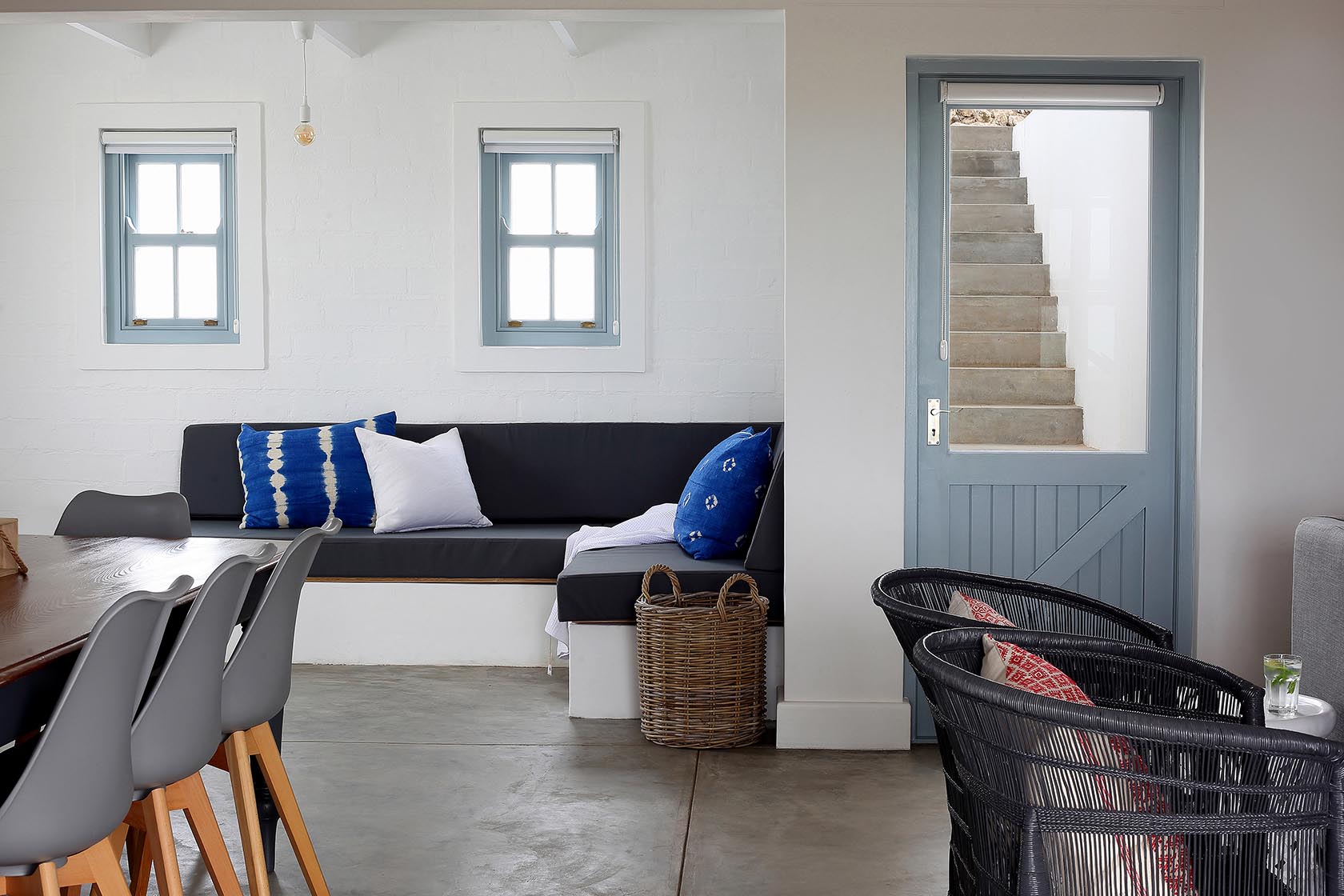 Perfect Hideaways – A bright, coastal-style interior with built-in charcoal seating, blue accent cushions, a dining table with modern grey chairs, and a view of exterior stairs through a blue door; Johanns Cottage, Churchhaven.