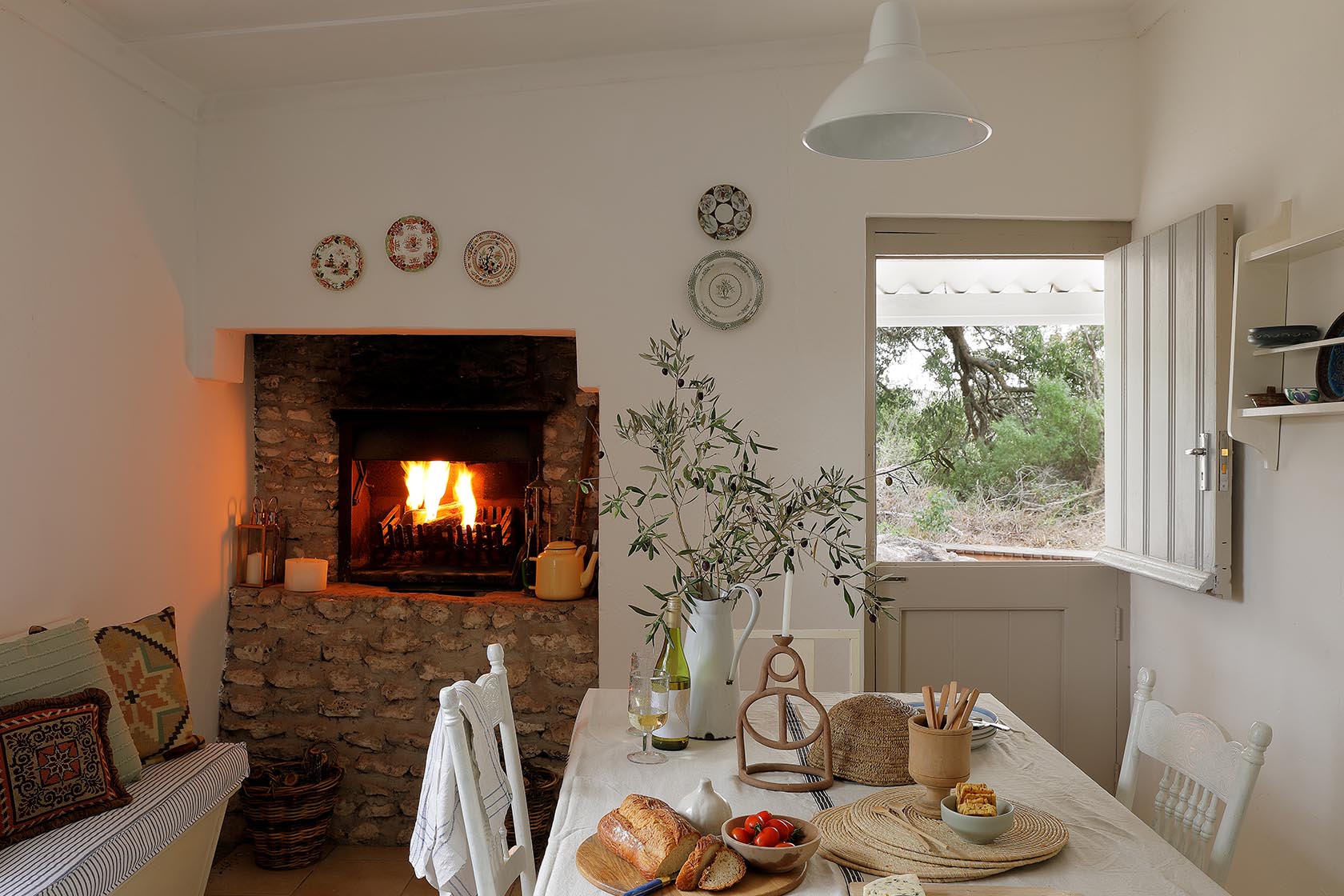 Perfect Hideaways. A rustic indoor dining area with a stone fireplace ablaze in the background, a set dining table with fresh bread and tomatoes, and a half-open stable door looking out to nature, Berit's Cottage, Churchhaven