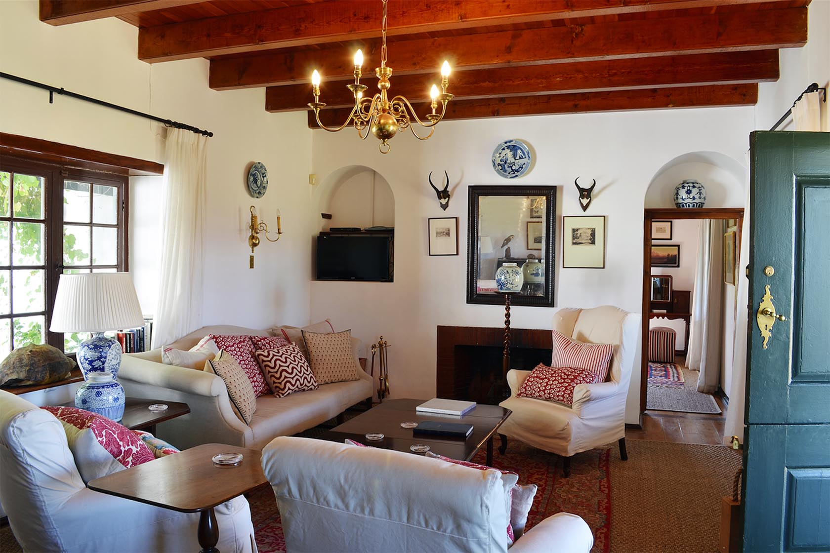 Perfect Hideaways – A cosy living room with exposed wooden beams, plush white armchairs, patterned cushions, antique decor, and a central fireplace creates a warm, traditional farmhouse atmosphere – Cottage 1685, Franschhoek, Western Cape, South Africa