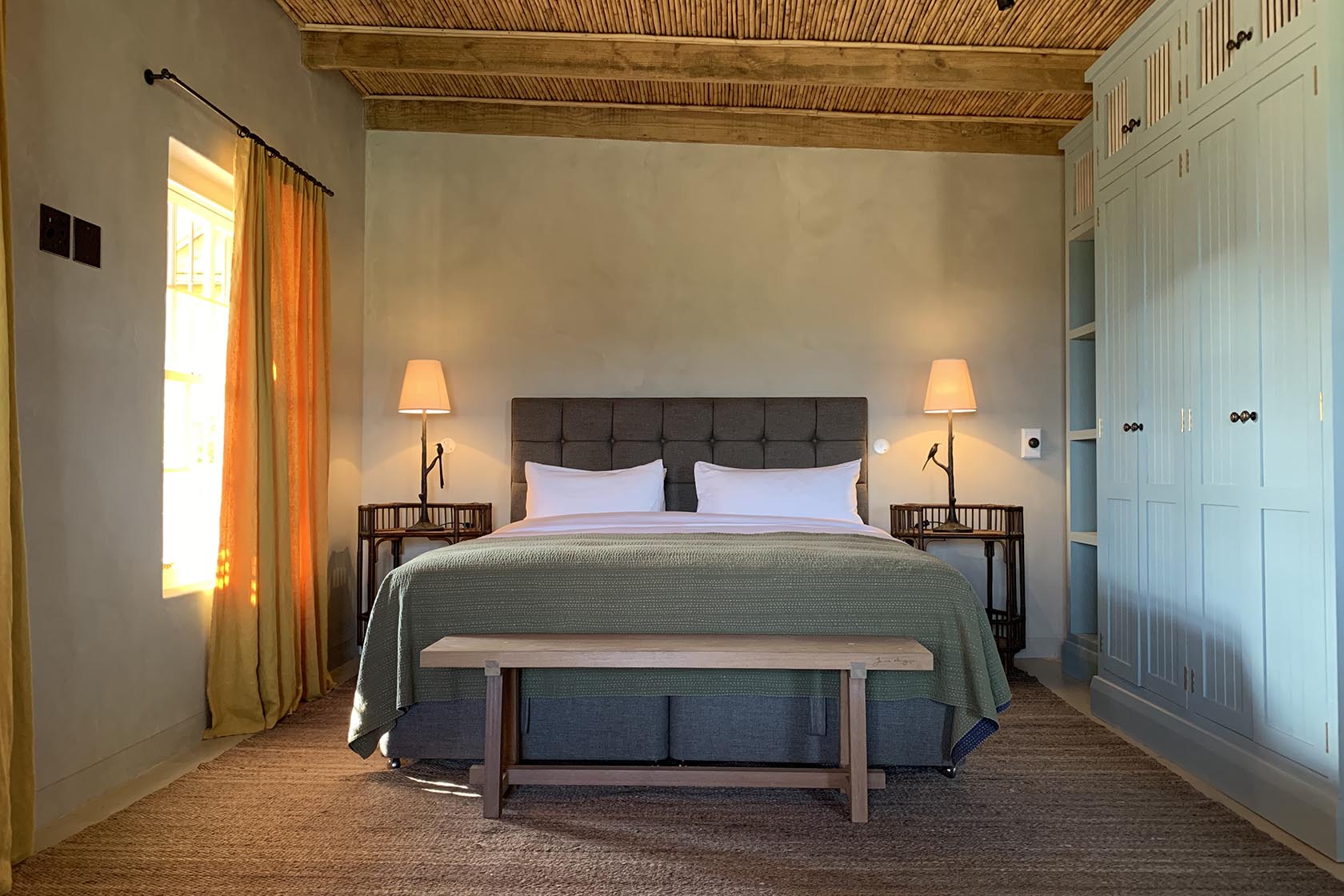 A serene bedroom with earthy tones, natural textures, a wooden ceiling, and soft light filtering through mustard curtains. Francois Cottage, Franschhoek