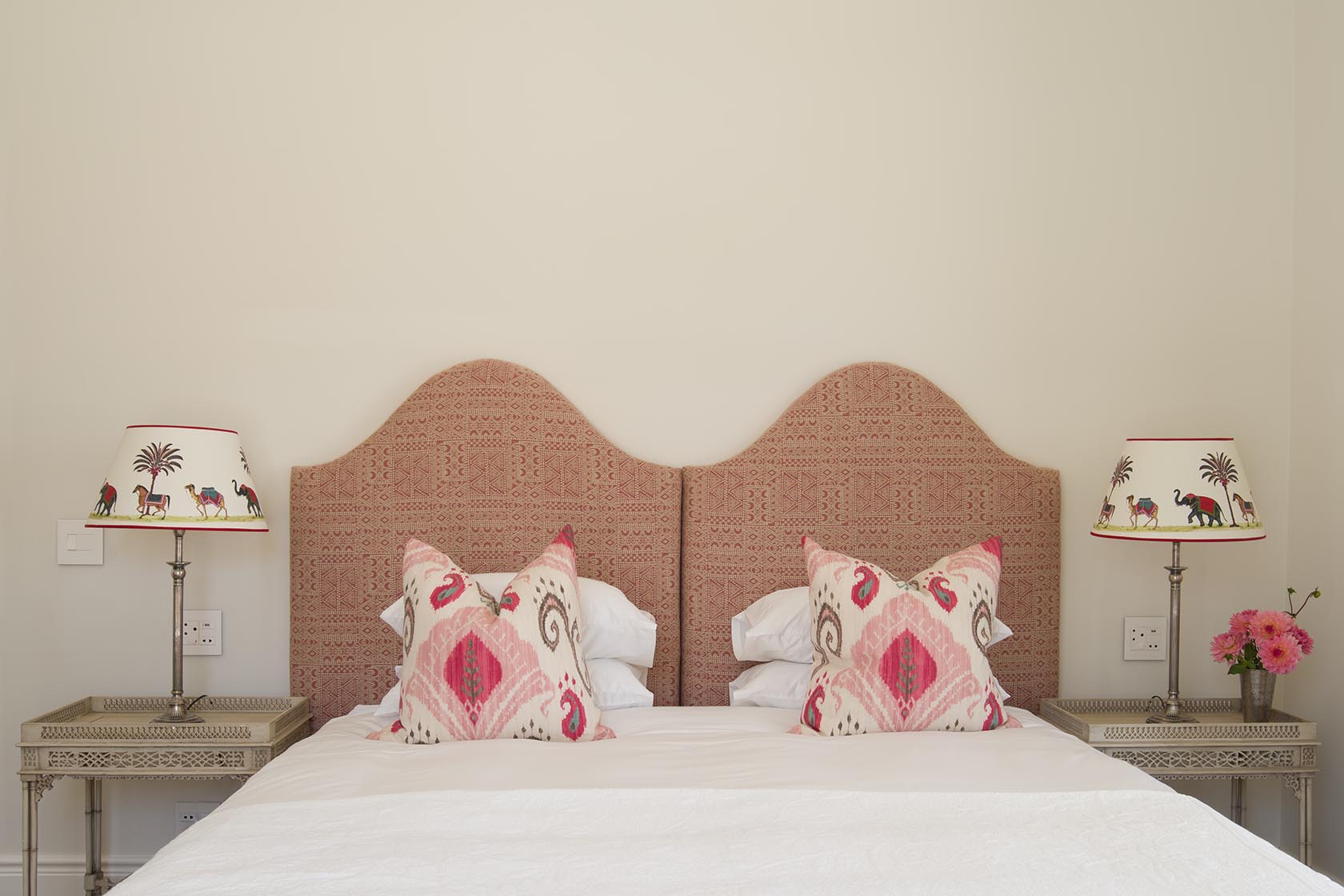 Perfect Hideaways – A bedroom decorated in warm pink and red tones, with a patterned twin headboard, pink and white accent pillows, silver bedside tables, and lampshades illustrated with camels and palm trees, creating a playful yet elegant atmosphere. La Cotte Orchard Cottages, Franschhoek South Africa