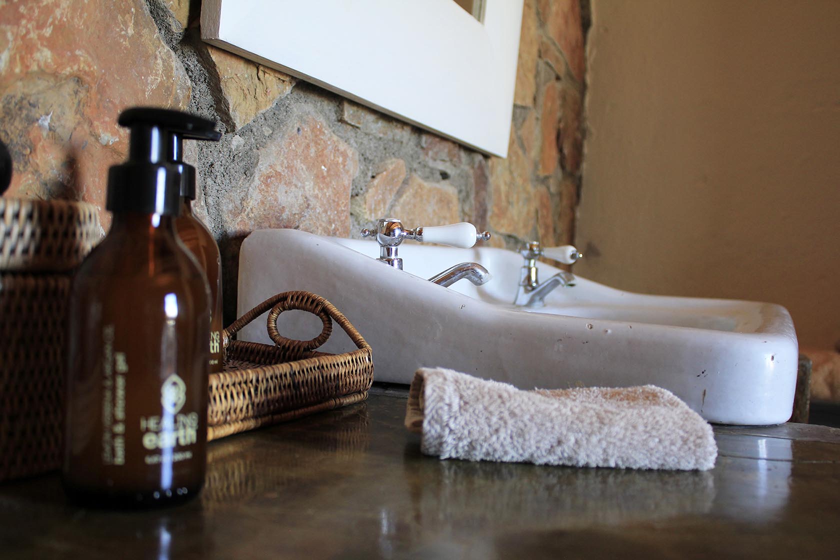 Perfect Hideaways- A close-up of a bathroom vanity with a white porcelain basin, chrome taps, woven baskets, amber glass toiletry bottles, and a soft folded towel set against a textured stone wall. Karoo Cottage, Sneeuberg Nature Reserve, Eastern Cape, South Africa