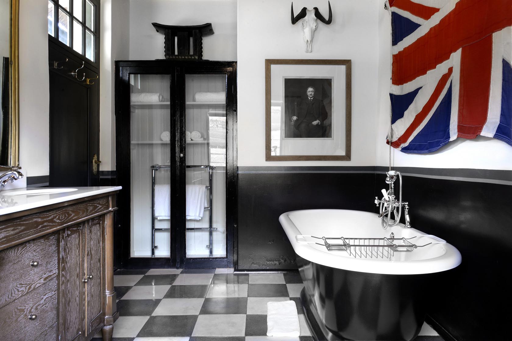 Perfect Hideaways – A dramatic black-and-white bathroom with a freestanding tub, vintage cabinetry, checkered floor, glass cabinet, an old portrait, and a large Union Jack flag hanging above – Cottage 1685, Franschhoek, Western Cape, South Africa