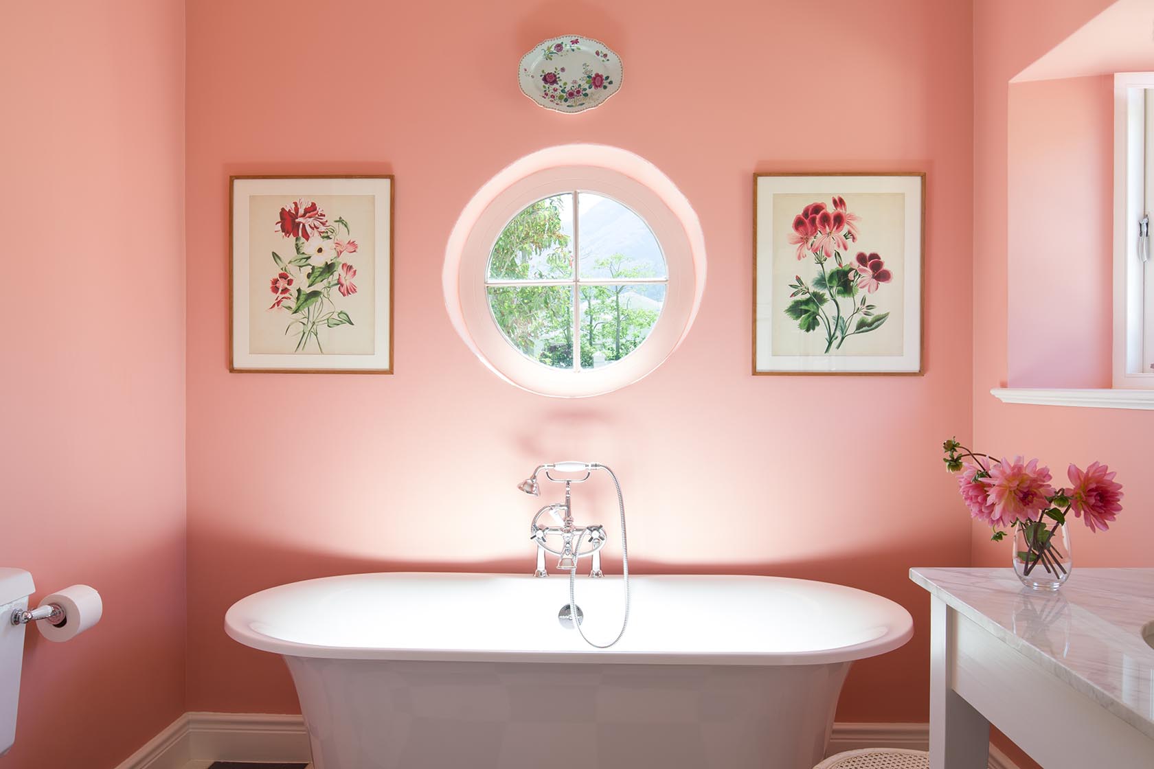 Perfect Hideaways – A soft pink bathroom with a freestanding tub beneath a round window, flanked by framed floral artworks, complemented by fresh pink flowers on the vanity and gentle natural light filling the room. La Cotte Orchard Cottages, Franschhoek South Africa