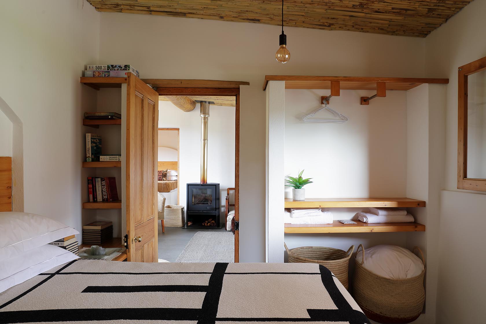 Perfect Hideaways – A bedroom with a geometric-patterned blanket, open shelving with baskets, and a peek into the living area where a fireplace glows in the distance. Soft natural tones and simple décor enhance the serene atmosphere. Sheperd's Cottage, Prince Albert