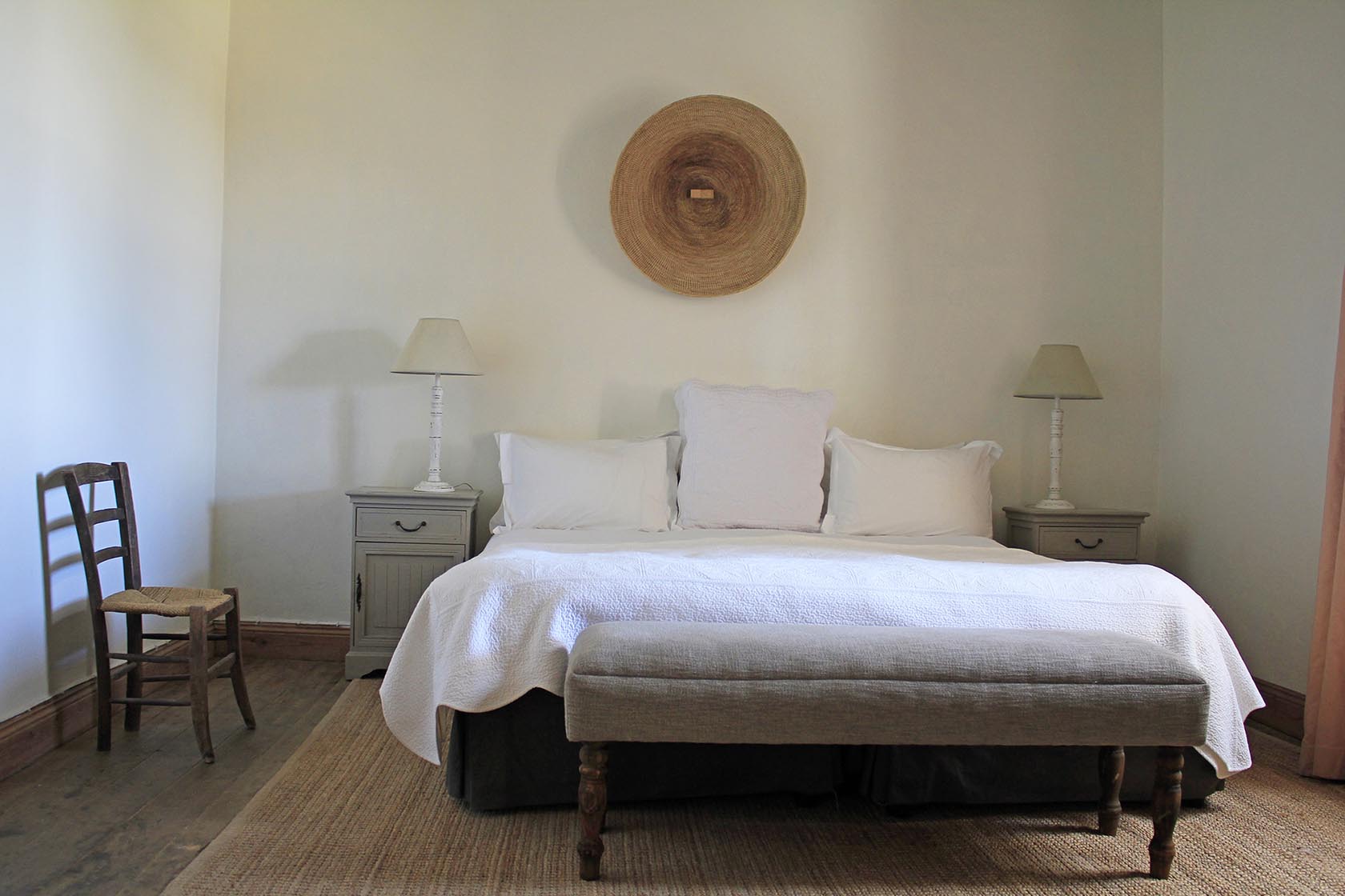 Perfect Hideaways- A serene bedroom with a large neatly made bed dressed in white linens, flanked by matching bedside tables with lamps, a woven rug underfoot, and a round woven wall piece centered above the headboard. Karoo Cottage, Sneeuberg Nature Reserve, Eastern Cape, South Africa