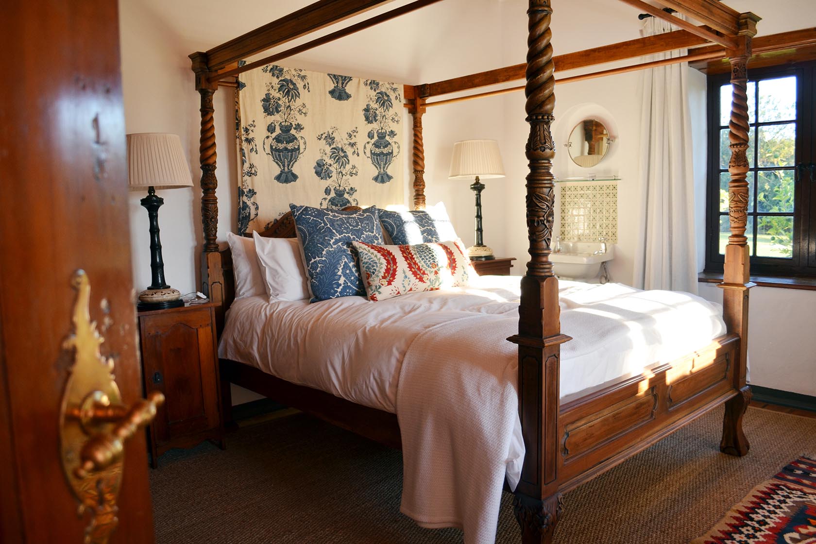 Perfect Hideaways – A warm, elegant bedroom with a carved wooden four-poster bed, patterned blue and red cushions, soft natural light, and antique bedside tables, creating a refined country ambiance – Cottage 1685, Franschhoek, Western Cape, South Africa
