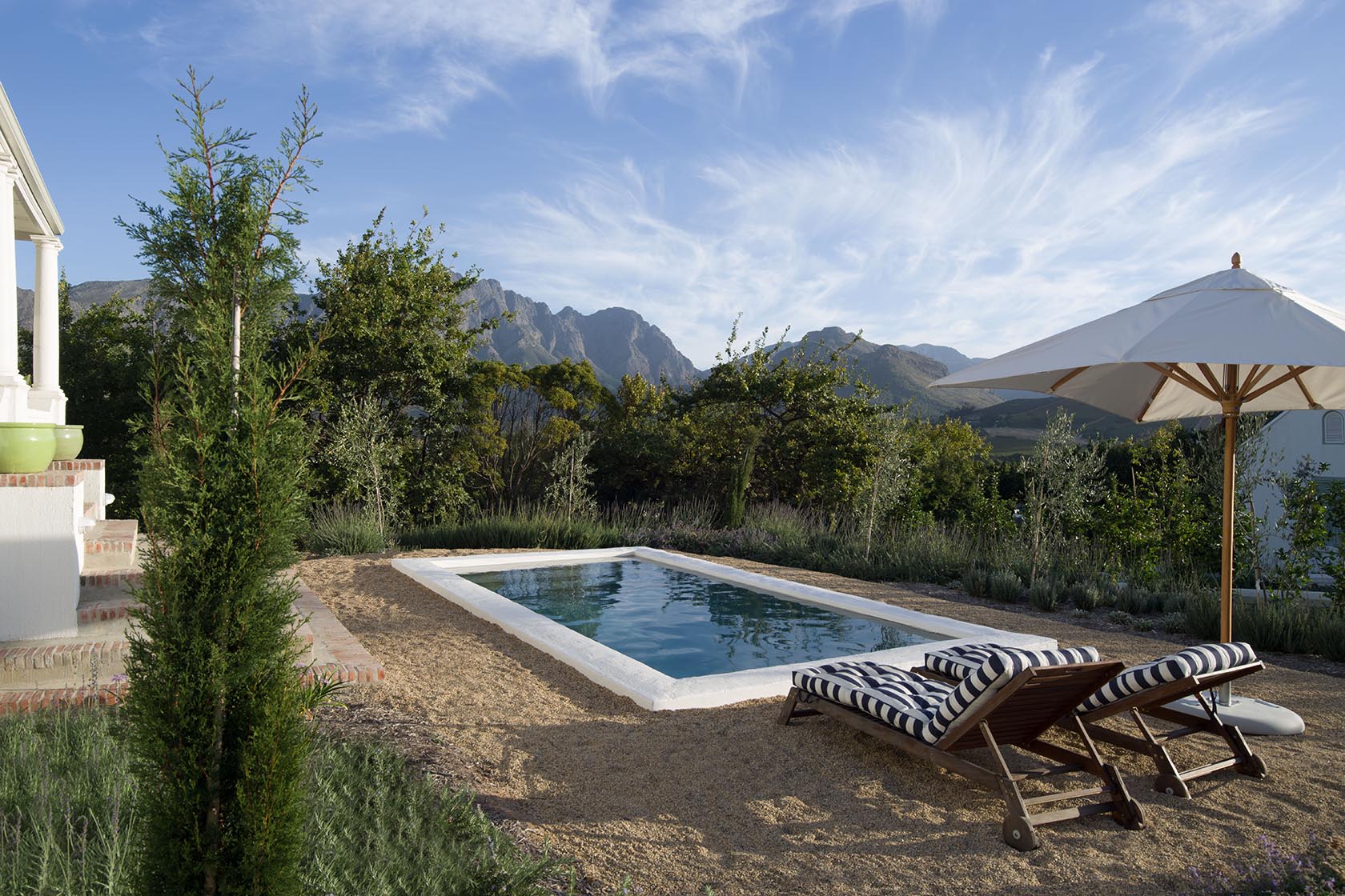 Perfect Hideaways – A tranquil outdoor pool surrounded by gravel and greenery, with striped loungers under a large white umbrella, set against sweeping mountain views and a bright blue sky with soft clouds. La Cotte Orchard Cottages, Franschhoek South Africa