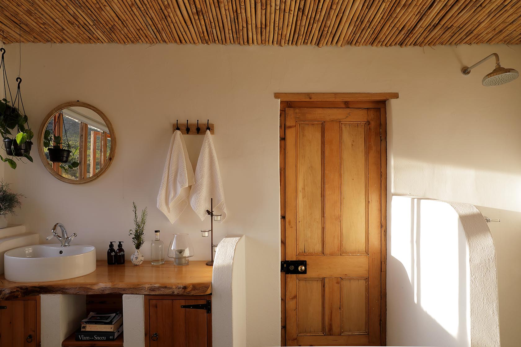 Perfect Hideaways – A bathroom with a bamboo ceiling, timber door, and a handcrafted wooden countertop supporting a round basin. Warm light filters in, illuminating hanging plants, glass bottles, and neatly hung white towels. Sheperd's Cottage, Prince Albert