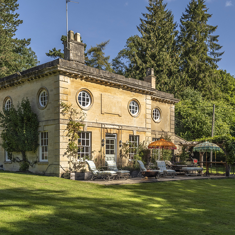Perfect Hideaways – A honey-coloured stone lodge with symmetrical round windows and classical architectural details, set within lush green lawns and tall surrounding trees. The patio features cushioned sun loungers, a firebowl, and vibrant parasols in orange and turquoise beneath a wooden pergola draped in greenery. Lince Lodge, England