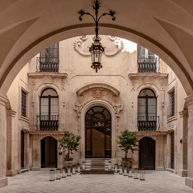 Perfect Hideaways – A grand, historic stone courtyard framed by high archways and ornate Baroque architectural details, featuring tall arched doorways, wrought-iron balconies, carved stone motifs, and two potted citrus trees flanking a central entrance, with rows of glass lanterns lining the stone floor – Palazzo Bozzi Corsa, Italy