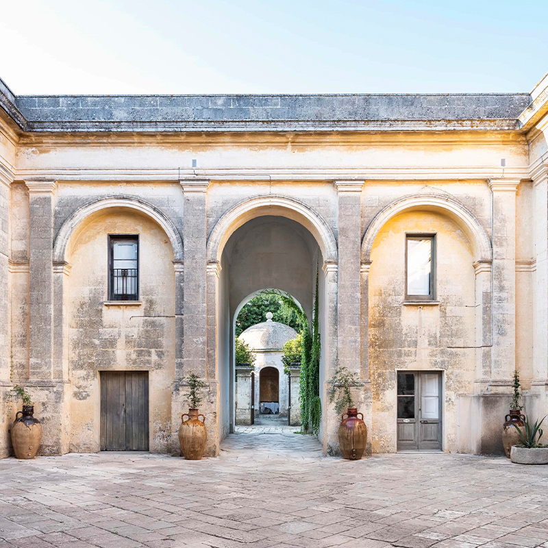 Desktop Banner 01. Perfect Hideaways – A grand historic courtyard framed by weathered stone arches and textured walls, featuring large earthenware vessels, rustic wooden doors, and a central arched passageway revealing a domed garden pavilion beyond, bathed in soft Mediterranean light – Palazzo Daniele_Salento_Puglia_Italy