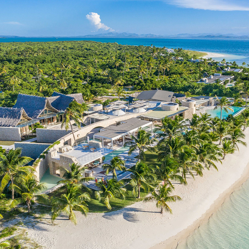 Perfect Hideaways – an aerial view of a secluded luxury island retreat surrounded by turquoise ocean, showing thatched-roof buildings, stone structures, lush palm trees, and a long stretch of pristine white-sand beach curving along the coastline, with villas tucked into dense tropical greenery and mountains faintly visible across the water in the distance – Miavana, Madagascar