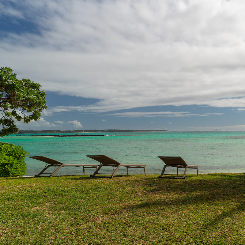 Perfect Hideaways – A tranquil beachfront scene with three sun loungers arranged on a grassy lawn overlooking a calm turquoise lagoon, framed by lush green foliage and a leaning palm tree under a soft, cloud-covered sky – Villa Saphir, Belle Mare