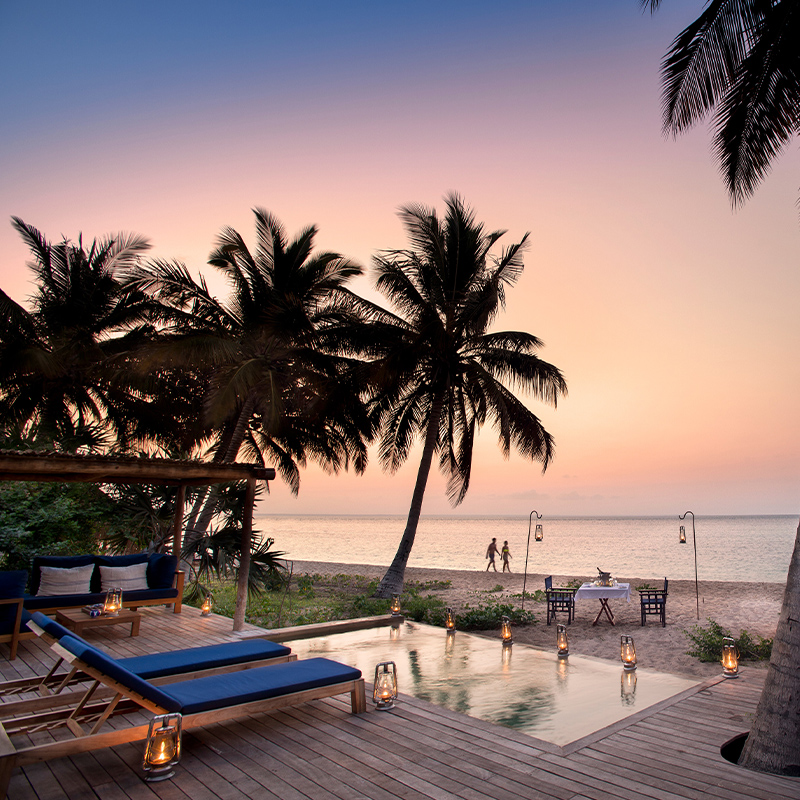 Perfect Hideaways - A serene beachfront deck at sunset featuring wooden loungers with blue cushions, a small reflective pool lined with lanterns, and views of palm trees framing a calm ocean where a couple walks along the shore; a private dining table is set on the sand and a thatched daybed sits nearby, all glowing in soft golden light. And Beyond Benguerra ,Mozambique