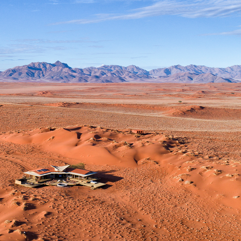 Perfect Hideaways - A panoramic aerial view of a luxury desert lodge built on raised wooden decks, featuring a circular plunge pool, shaded outdoor lounges, and solar-panel rooftops, set among sweeping red sand dunes with distant mountain ranges under a bright blue sky. Mountain View Suite, NamibRand Nature reserve