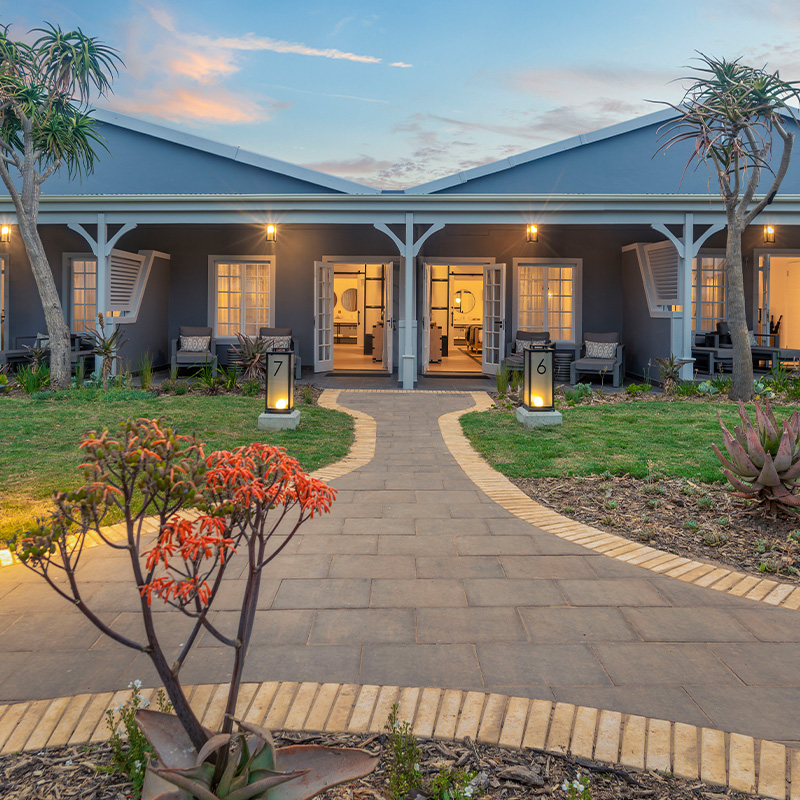 Perfect Hideaways – A twilight exterior view of two adjoining guest suites with open French doors glowing warmly from inside, framed by soft grey walls, white trim, and a shaded verandah. A curved garden pathway leads through neatly landscaped greenery with aloes and succulents toward the entrances marked “6” and “7,” while tall aloe trees stand on either side beneath a pastel sunset sky. Riverdene, Shamwari Private Game Reserve, Eastern Cape, South Africa