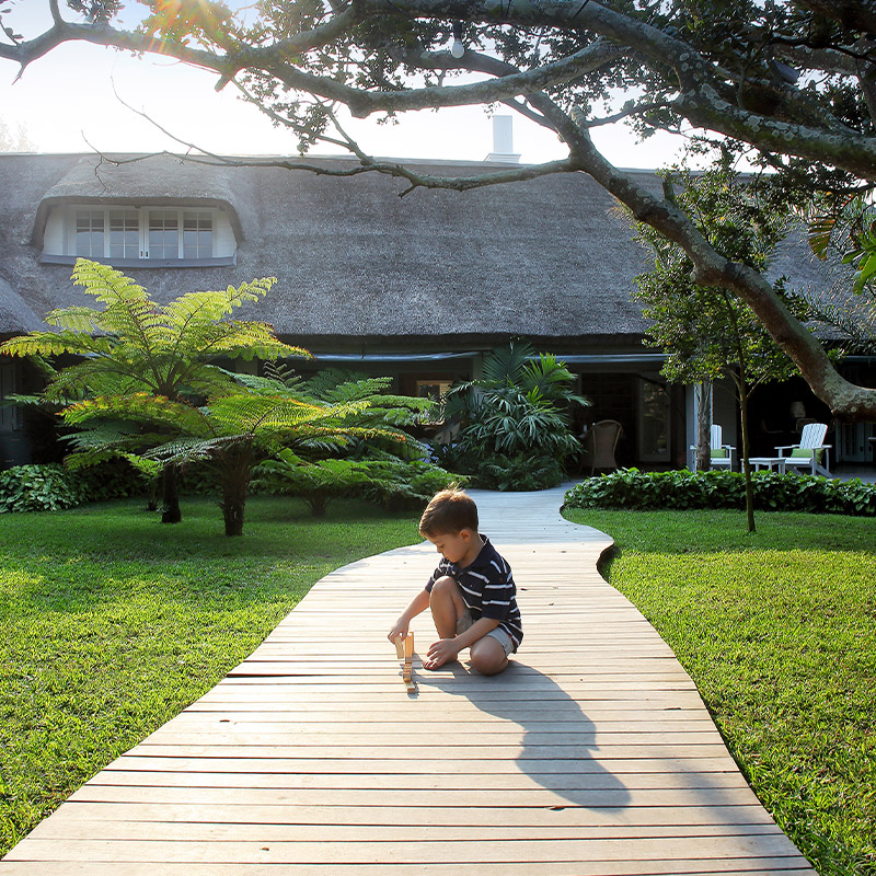 Perfect Hideaways- A young child sits and plays on a wooden walkway winding through a lush green garden, leading toward a thatched-roof cottage shaded by trees and tropical plants cowrie Cottage, Dolphin Coast, Kwazulu- Natal, South Africa