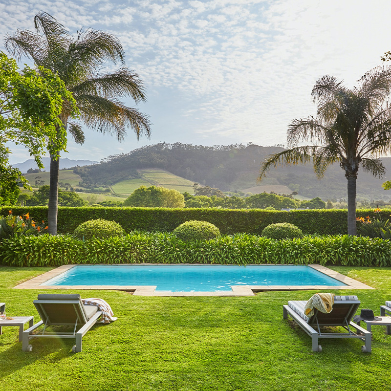 Perfect Hideaways: A serene swimming pool surrounded by neatly manicured lawns, palm trees, and vibrant greenery, with rolling vineyards and mountains in the background. Key To The Mountains, Franschhoek.