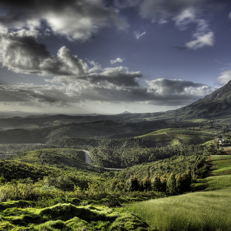 Perfect Hideaways- panoramic view of rolling green vineyards, winding roads, and layered mountain ranges under dramatic cloud-filled skies, with sunlight casting soft highlights across the valley, Delaire Graff, Stellenbosch, Western Cape, South Africa