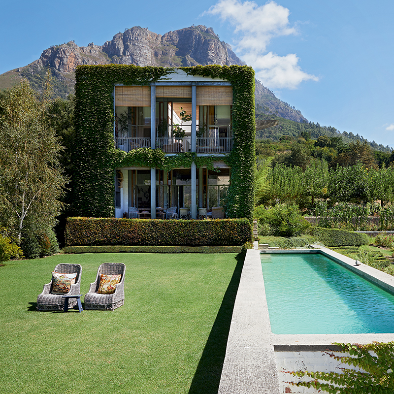 Perfect Hideaways: A stunning two-storey home covered in green ivy, set against a backdrop of majestic mountains, with a pristine swimming pool and manicured lawns in the foreground. Johannesdal Manor, Stellenbosch.