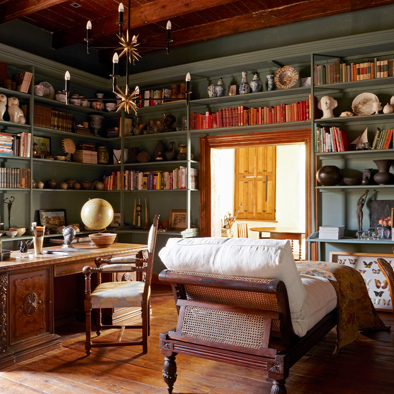 Perfect Hideaways- A character-filled study with floor-to-ceiling sage-green bookshelves lined with books, ceramics, sculptures and curios, a carved wooden desk with a globe and writing chair, a cane-backed daybed, warm timber floors and starburst pendant lights, with sunlight streaming through an open doorway Jonkmanshof, Montague, Western Cape, South Africa