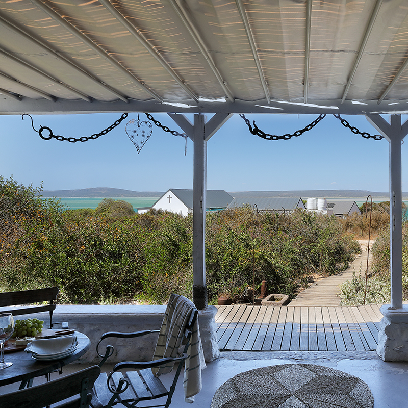 Perfect Hideaways–View from a shaded veranda across coastal fynbos toward a turquoise lagoon, with a white chapel and cottages in the distance and an outdoor table set for dining in the foreground. Church Cottage, Churchhaven, South Africa