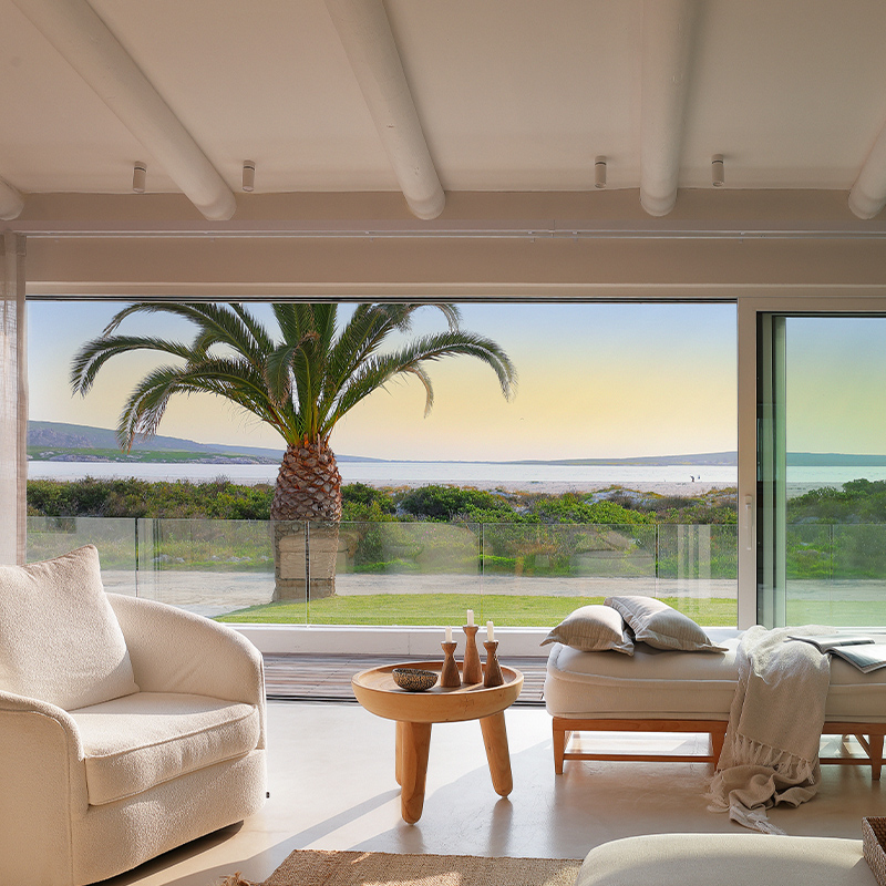 Perfect Hideaways – A serene, light-filled living space with soft neutral furnishings, including a curved armchair, wooden side table with sculptural candle holders, and a cushioned daybed draped with a throw, all set before expansive floor-to-ceiling glass doors that frame a tranquil coastal view with a tall palm tree, greenery, and calm water under a pastel sky – Kinsail Beach Villa, Langebaan