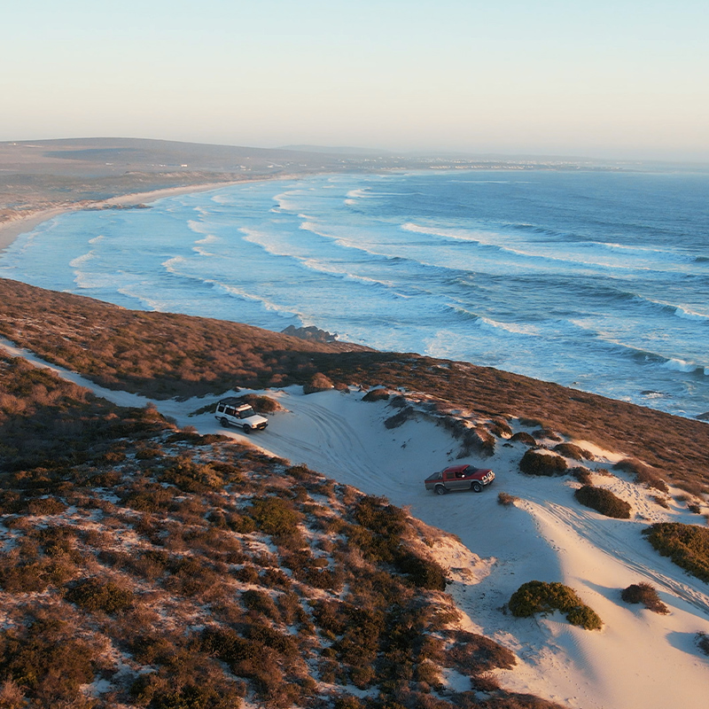 Perfect Hideaways – An aerial view of a rugged West Coast shoreline with golden, scrub-covered dunes meeting a curve of sandy beach. A winding dirt track leads toward the ocean, where dark rocks break the surf and waves roll in under soft, warm light. Treskoskraal Farm, Parternoster